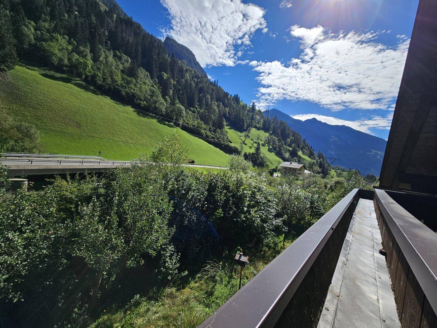 Weitläufiger Blick von einem Balkon auf eine grüne Berglandschaft und eine Straße.