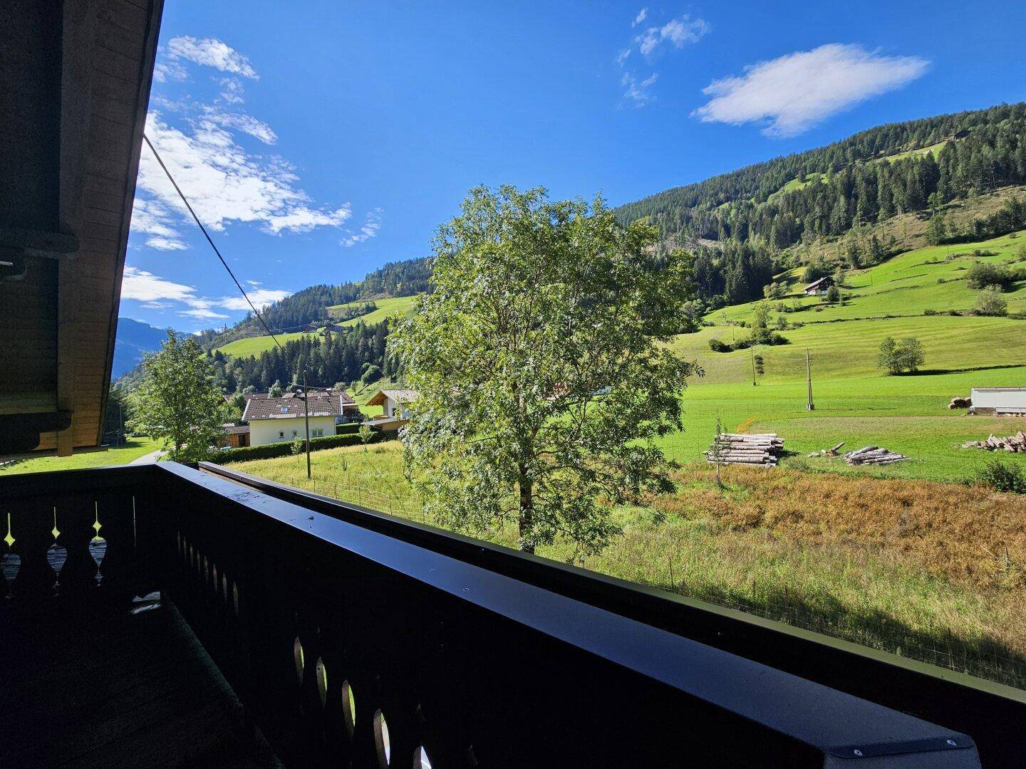 Panoramablick von einem Balkon über grüne Wiesen und bewaldete Berge unter blauem Himmel.
