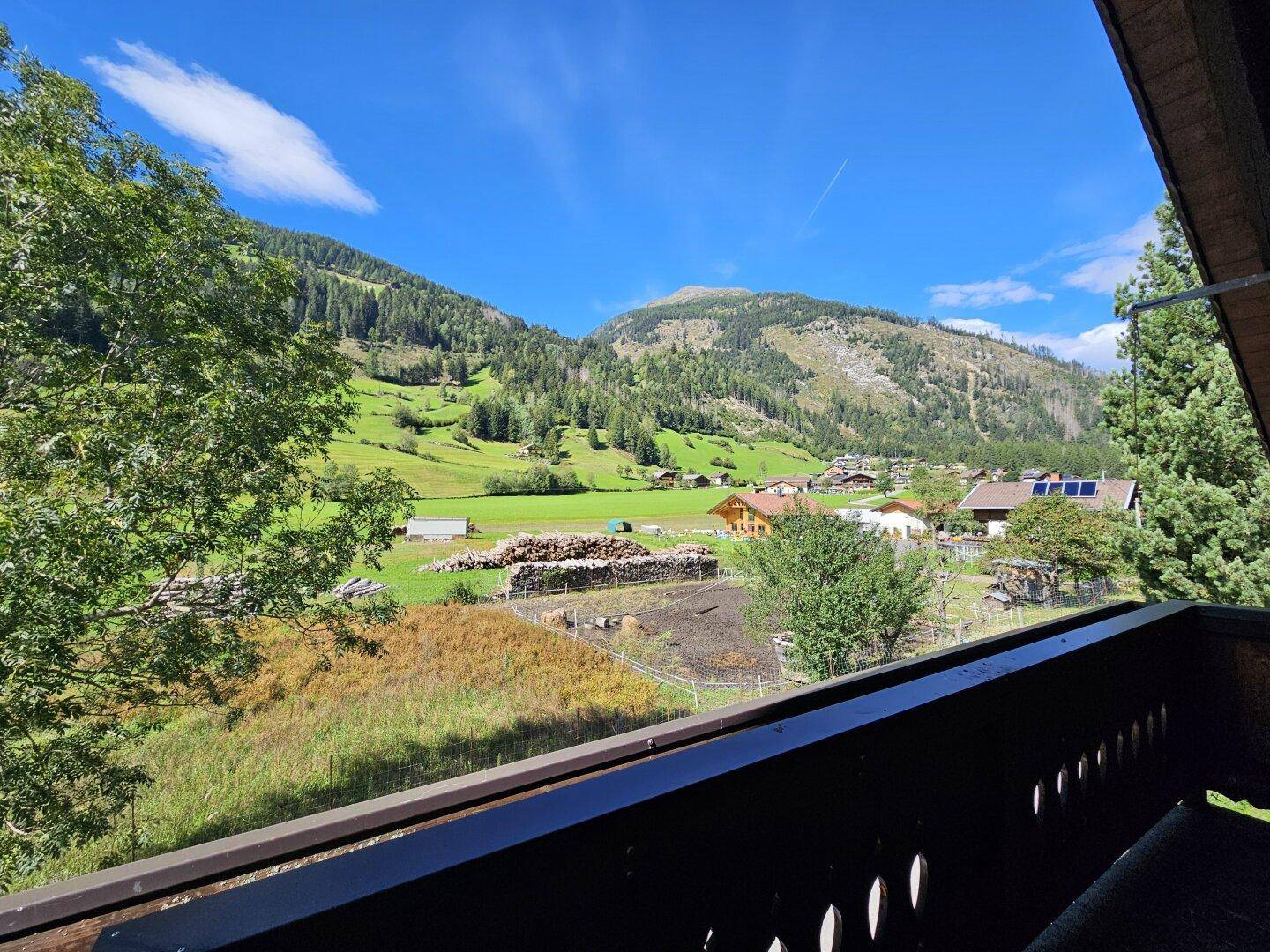 Blick vom Balkon auf eine malerische Berglandschaft mit grünen Wiesen und einem kleinen Dorf unter blauem Himmel.