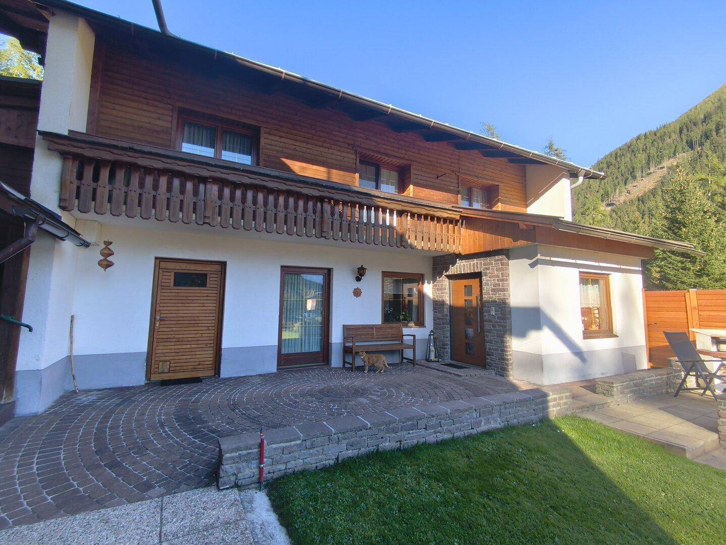 Charmante Hausfassade mit Holzverkleidung, einem gepflasterten Terrassenbereich und einem Holzbalkon.