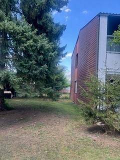 Seitenansicht des Gebäudes mit dunkler Fassade, umgeben von hohen Bäumen und einer Rasenfläche.
