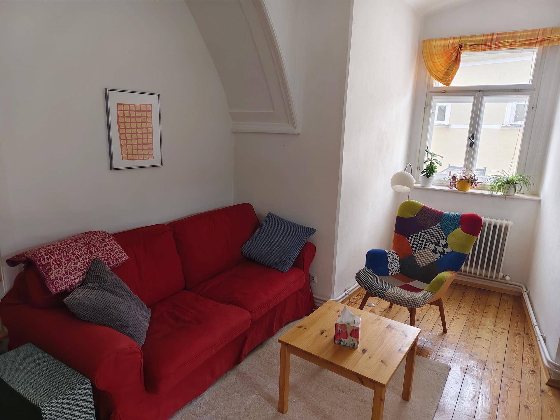 Wohnzimmer mit rotem Sofa, farbenfrohem Sessel und Holzboden, hell durch großes Fenster.