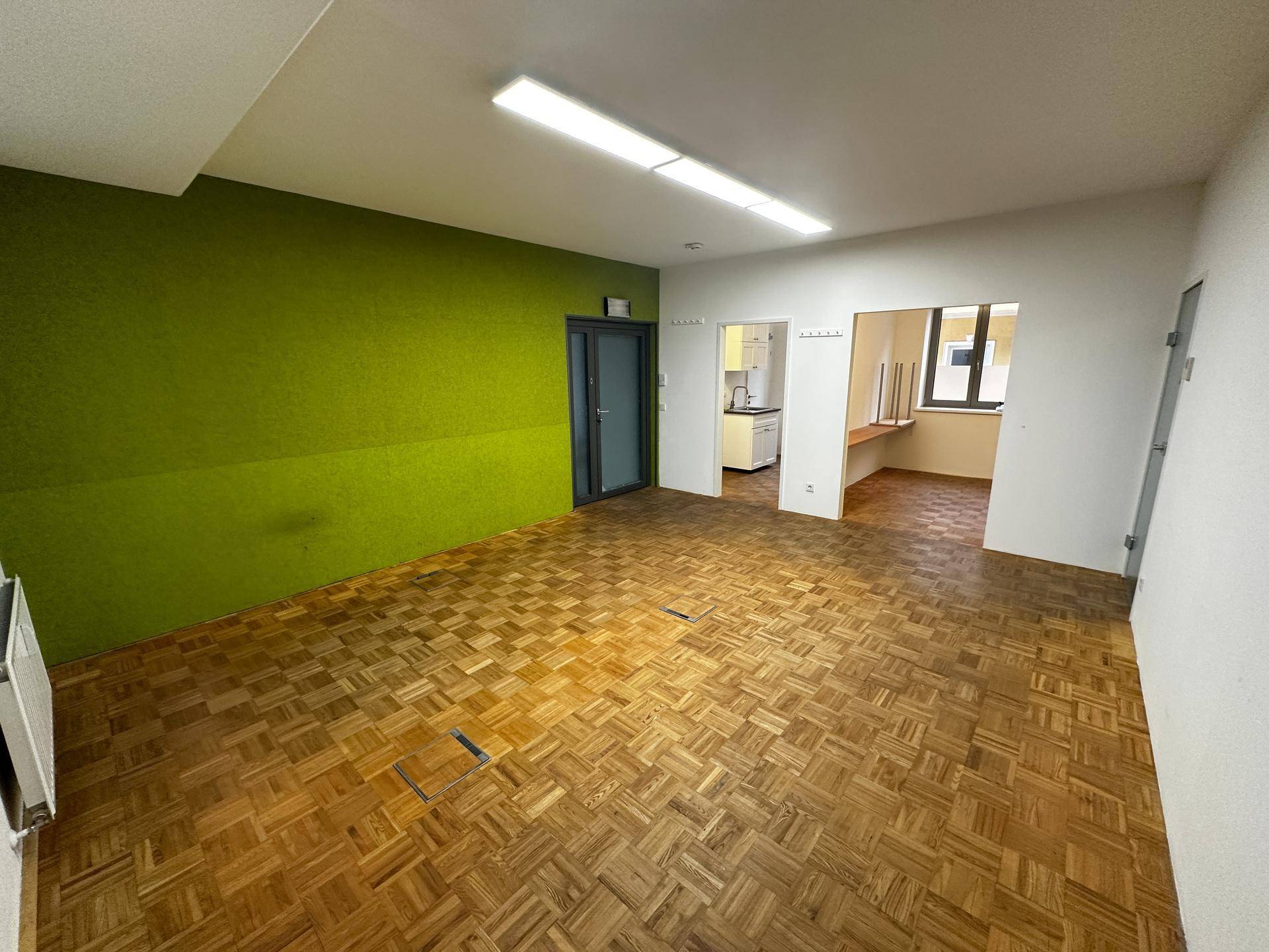Großer, offener Bürobereich mit Holzparkett, einer grünen Akzentwand und Zugang zu weiteren Räumen.
