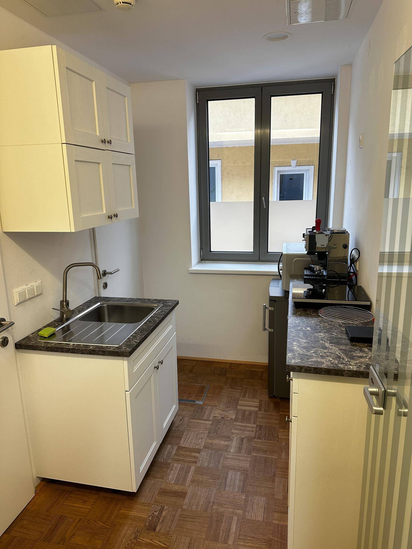 Kleine Küchenzeile mit weißen Schränken, dunkler Arbeitsplatte und Fenster mit Blick auf den Innenhof.
