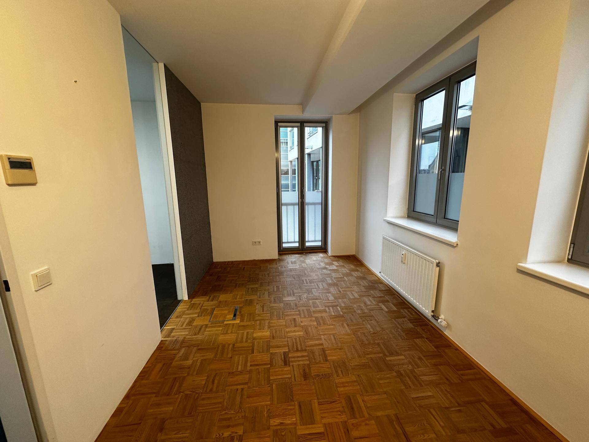 Heller Raum mit Fischgrätparkett, großen Fenstern und Zugang zu einem Balkon oder einer Loggia.