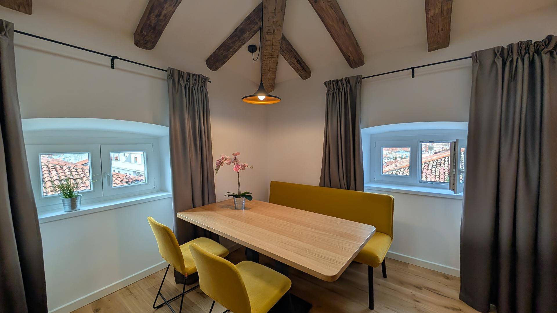 Gemütlicher Essbereich mit Holztisch, gelber Sitzbank und Fenstern mit Blick auf die Dächer.