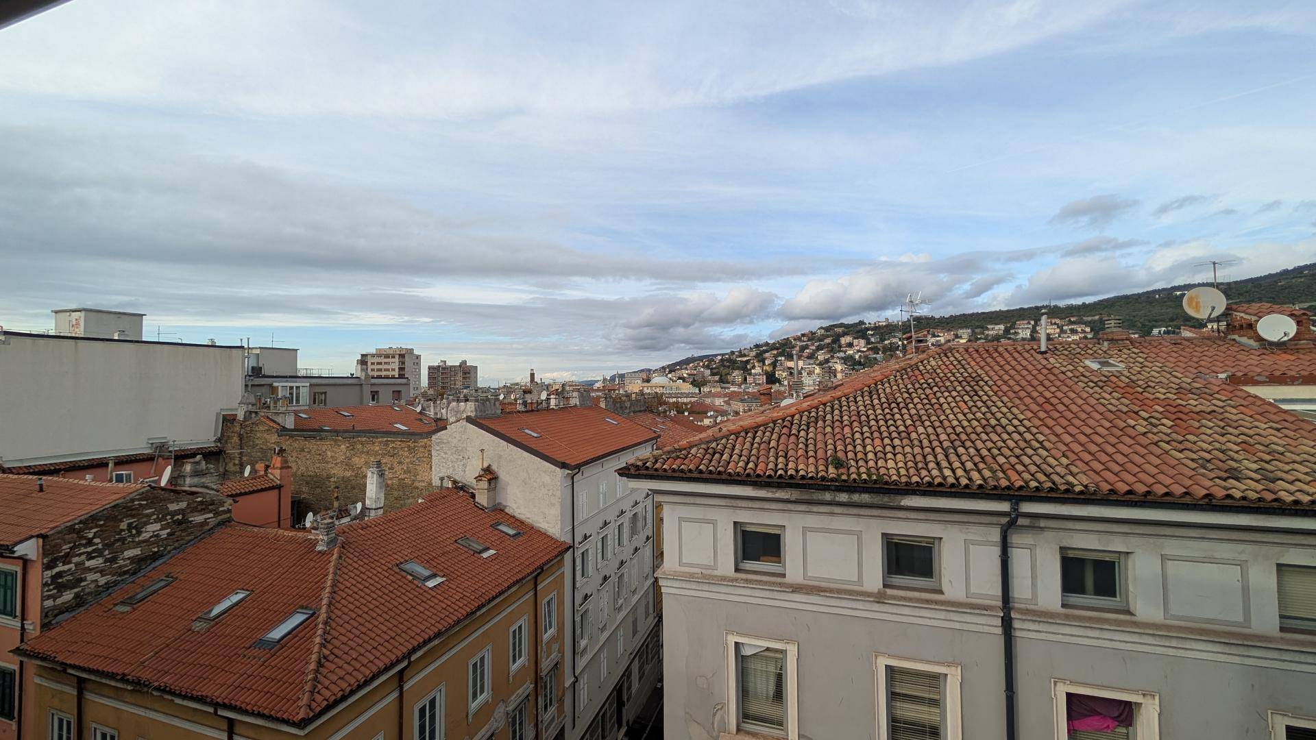 Panoramablick über die Dächer einer Stadt mit Hügeln im Hintergrund unter bewölktem Himmel.