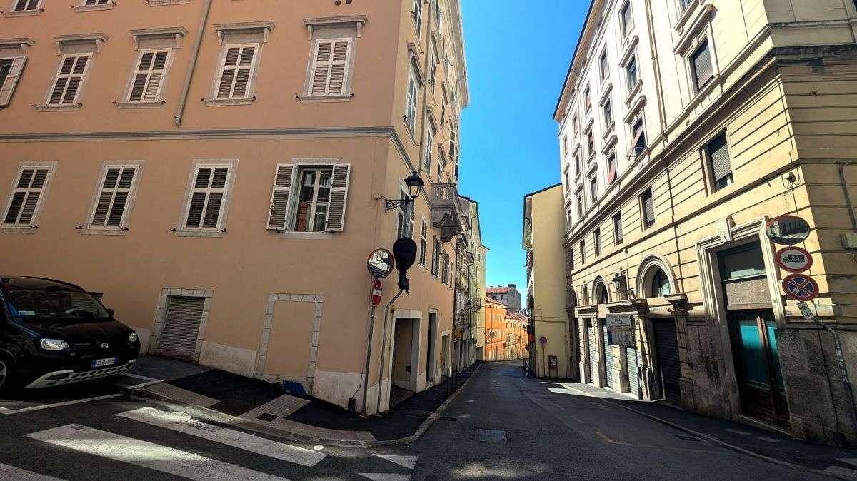 Enge Gasse in einer historischen Stadt mit geparkten Autos und alten Gebäuden unter blauem Himmel.