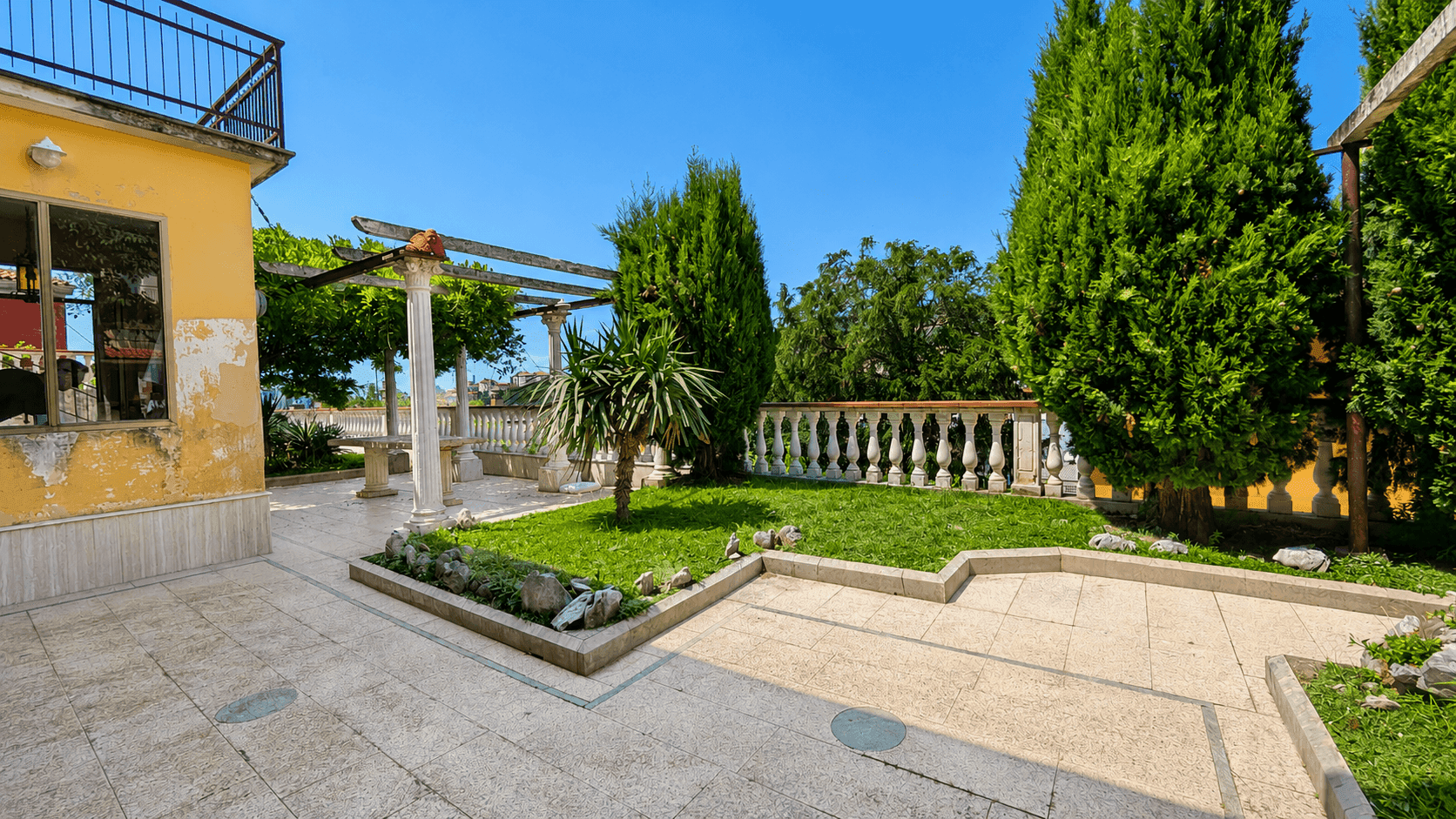 Gepflegter Gartenbereich mit Rasenflächen, gepflasterten Wegen und Zypressen unter strahlend blauem Himmel.