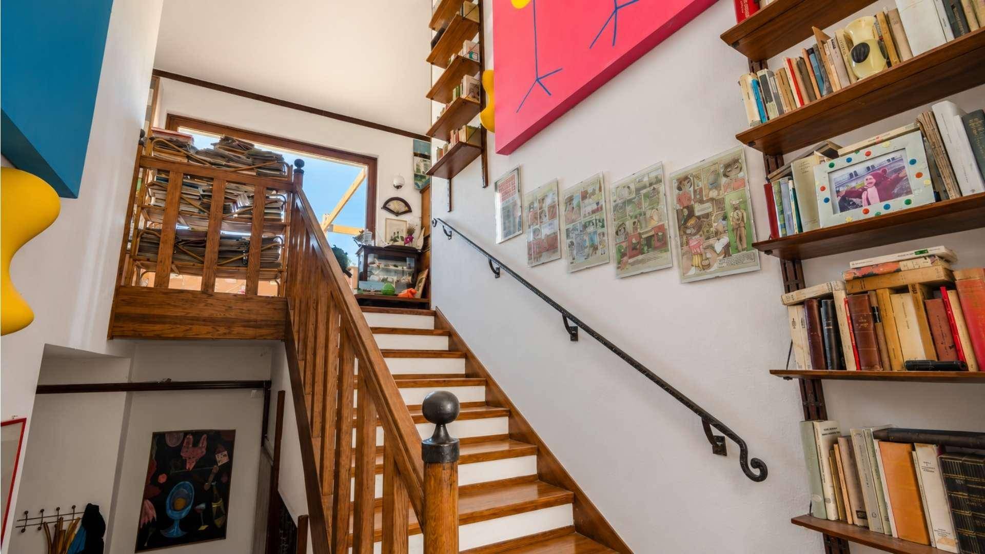 Treppenhaus mit Holzgeländer, Bücherregalen und bunten Kunstwerken an den Wänden.