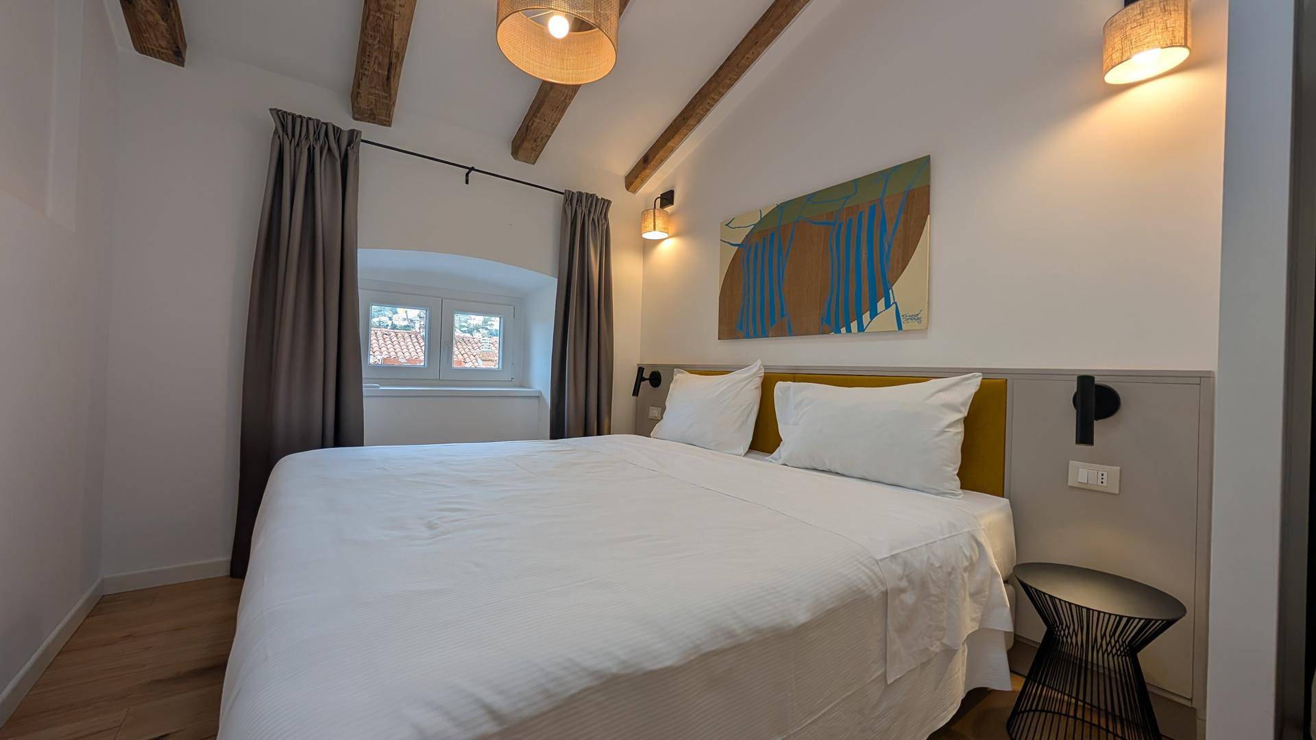 Gemütliches Schlafzimmer mit Doppelbett, Holzbalken und Fenstern mit Blick auf die Dächer.