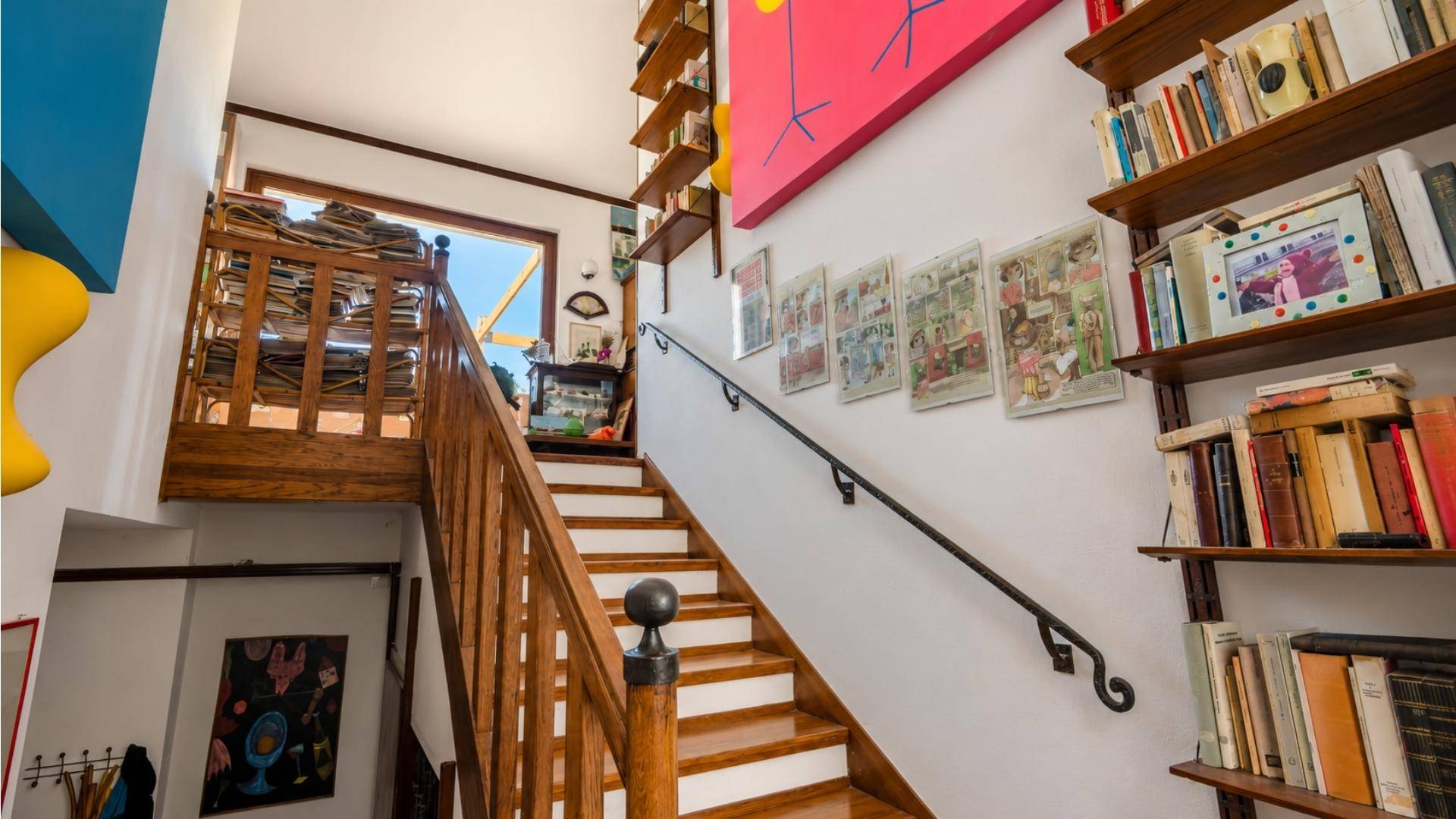 Holztreppe mit Geländer und farbenfrohen Kunstwerken an den Wänden, die nach oben führt.