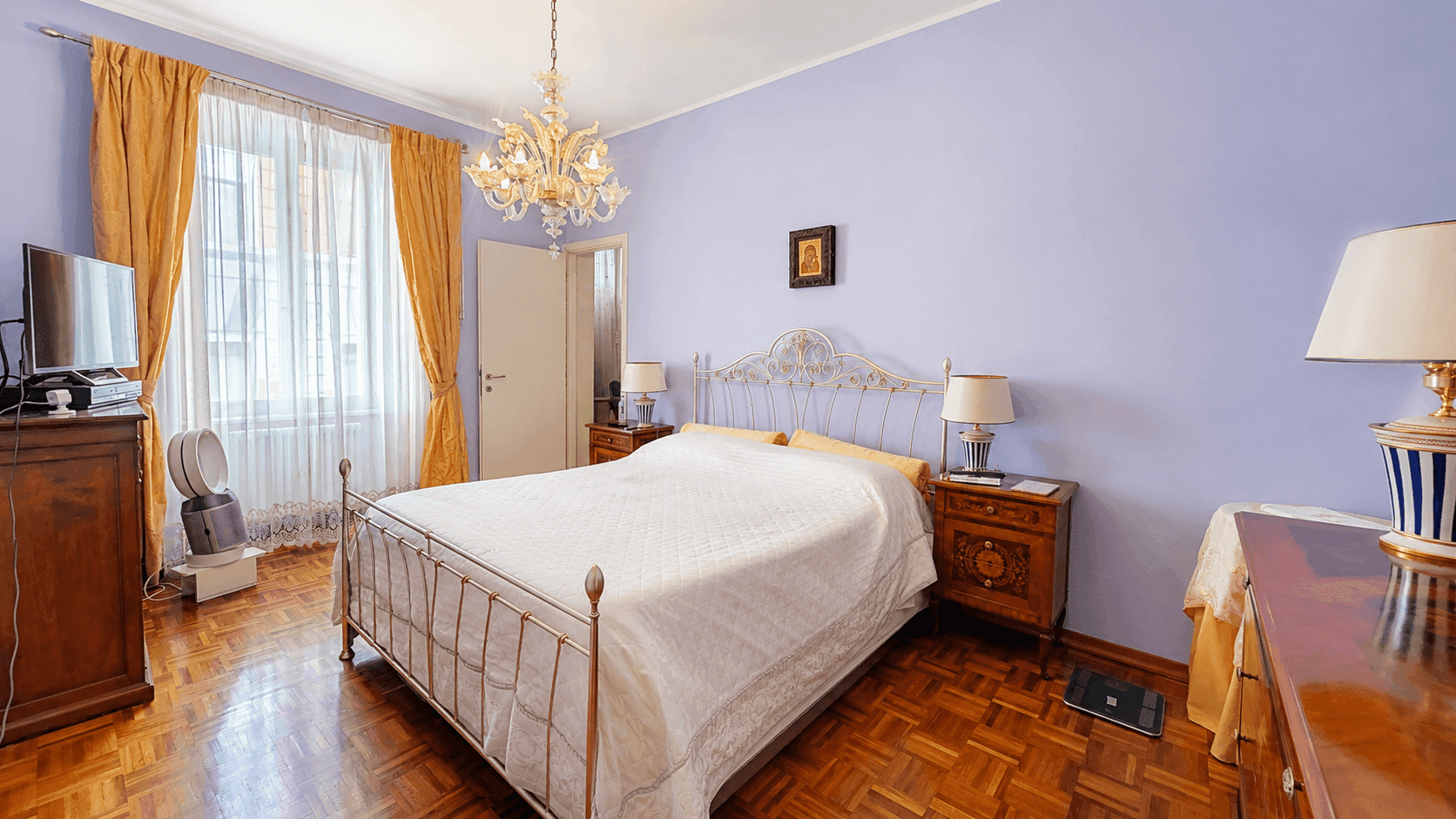Schlafzimmer mit lila Wänden, Parkettboden, Doppelbett und einem eleganten Kronleuchter, hell durch große Fenster.