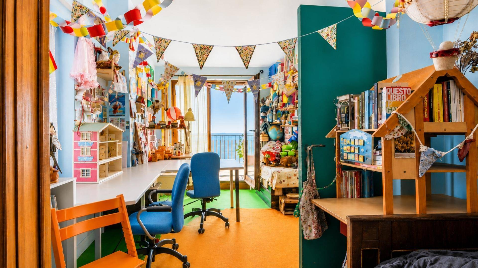 Kreativ gestaltetes Zimmer mit bunten Girlanden, Schreibtisch und Zugang zu einem Außenbereich.