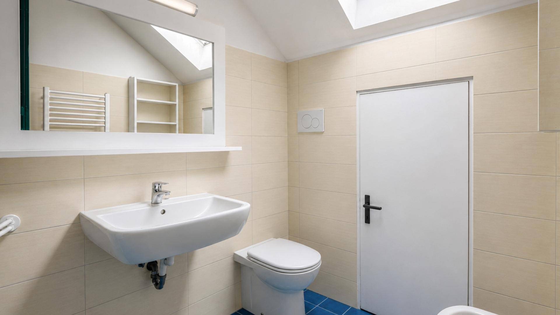 Modernes Badezimmer mit beigen Fliesen, Waschbecken, Toilette und Oberlicht für Tageslicht.