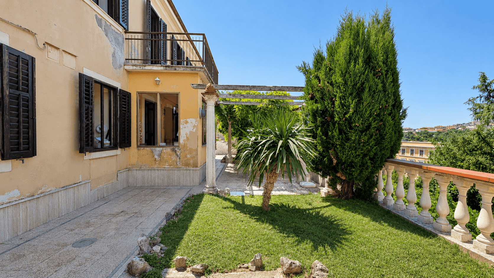 Sonniger Gartenbereich neben dem Haus mit Rasen, Palmen und einer Pergola, umgeben von einer Balustrade.