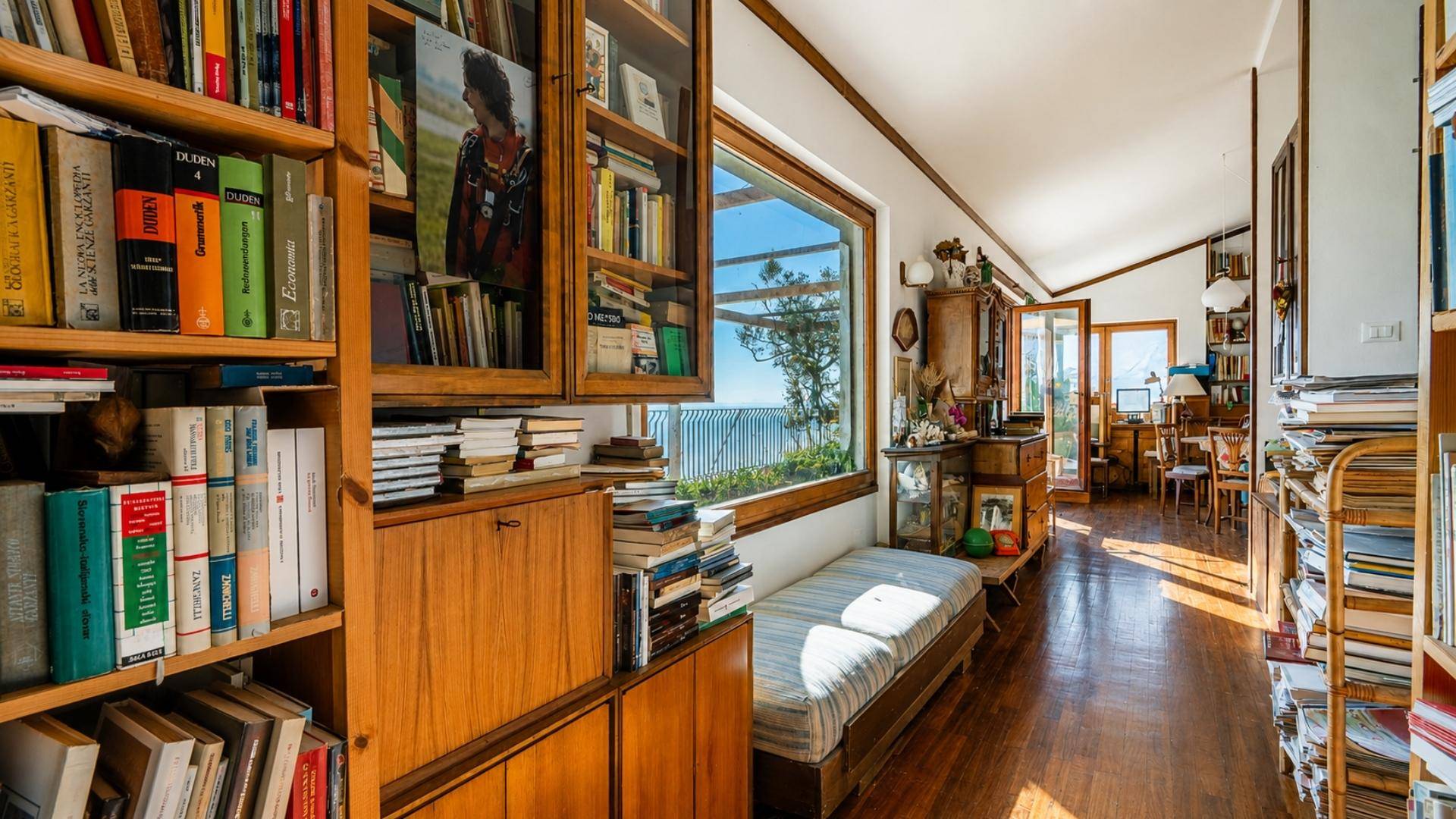 Heller offener Bereich mit Holzboden, Bücherregalen und seitlichem Meerblick durch ein großes Fenster.