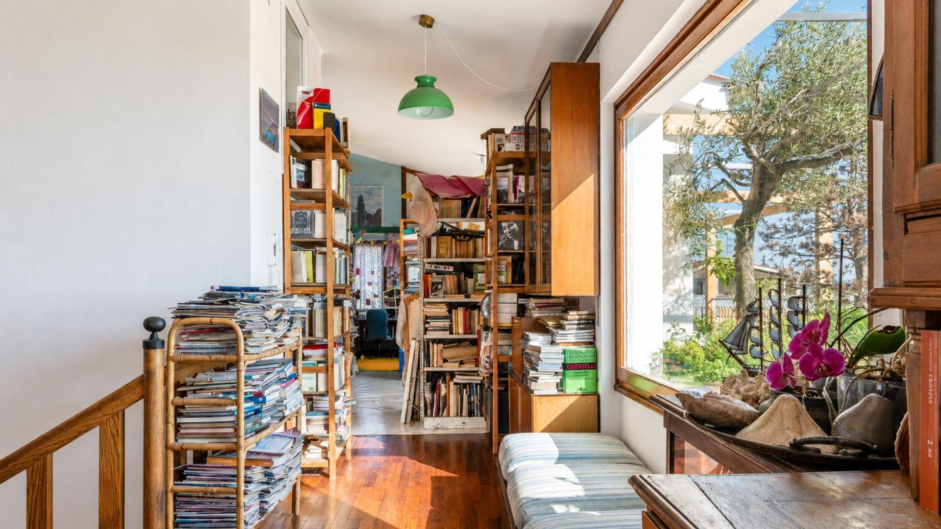 Treppenbereich mit Holzgeländer und Bücherregalen, hell durch ein großes Fenster mit Grünblick.