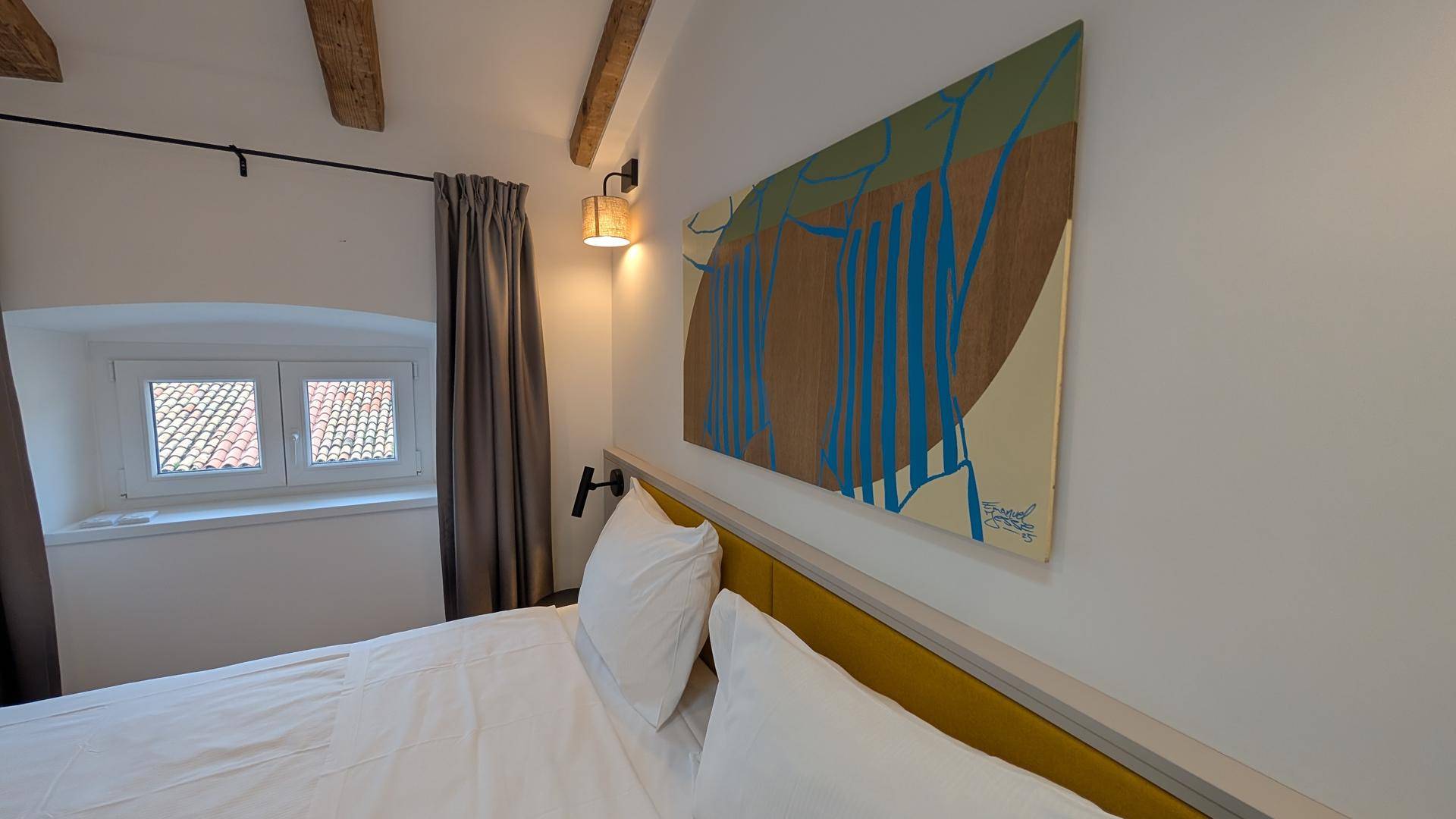 Schlafzimmer mit Doppelbett, modernem Kunstwerk und Fenstern mit Blick auf die Dächer.