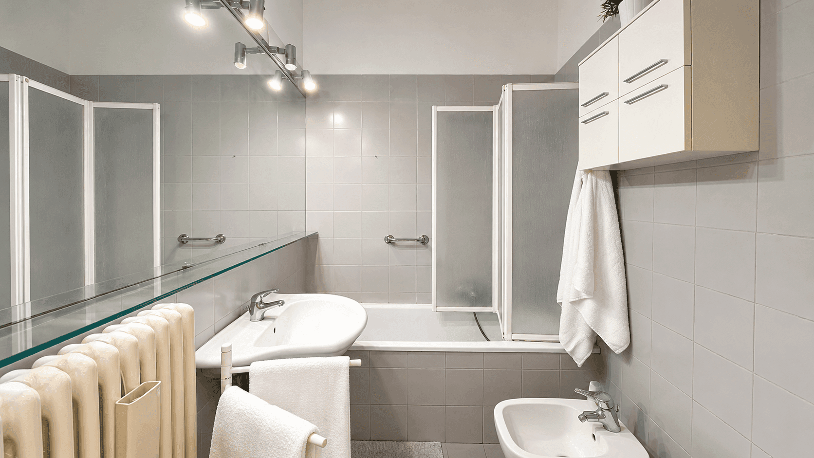 Funktionales Badezimmer mit grauen Fliesen, Badewanne mit Duschabtrennung und Bidet, ausgestattet mit Haltegriffen.