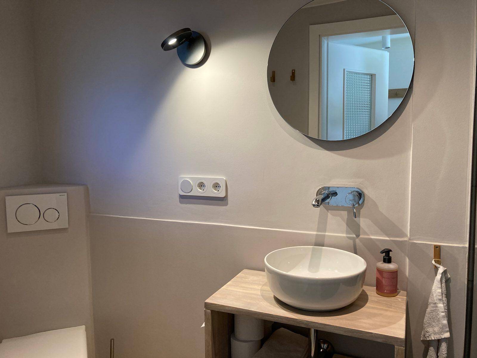 Modernes Badezimmer mit Aufsatzwaschbecken, rundem Spiegel und Wand-WC in hellen Farbtönen.