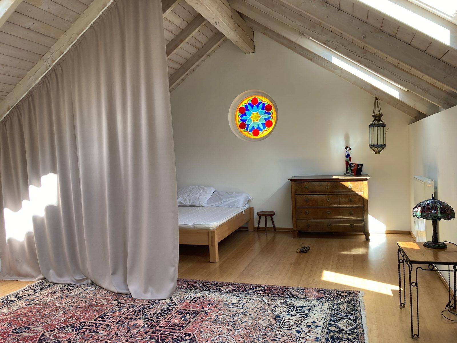 Gemütliches Schlafzimmer im Dachgeschoss mit Holzbalken, schrägen Wänden und einem farbigen Rundfenster.