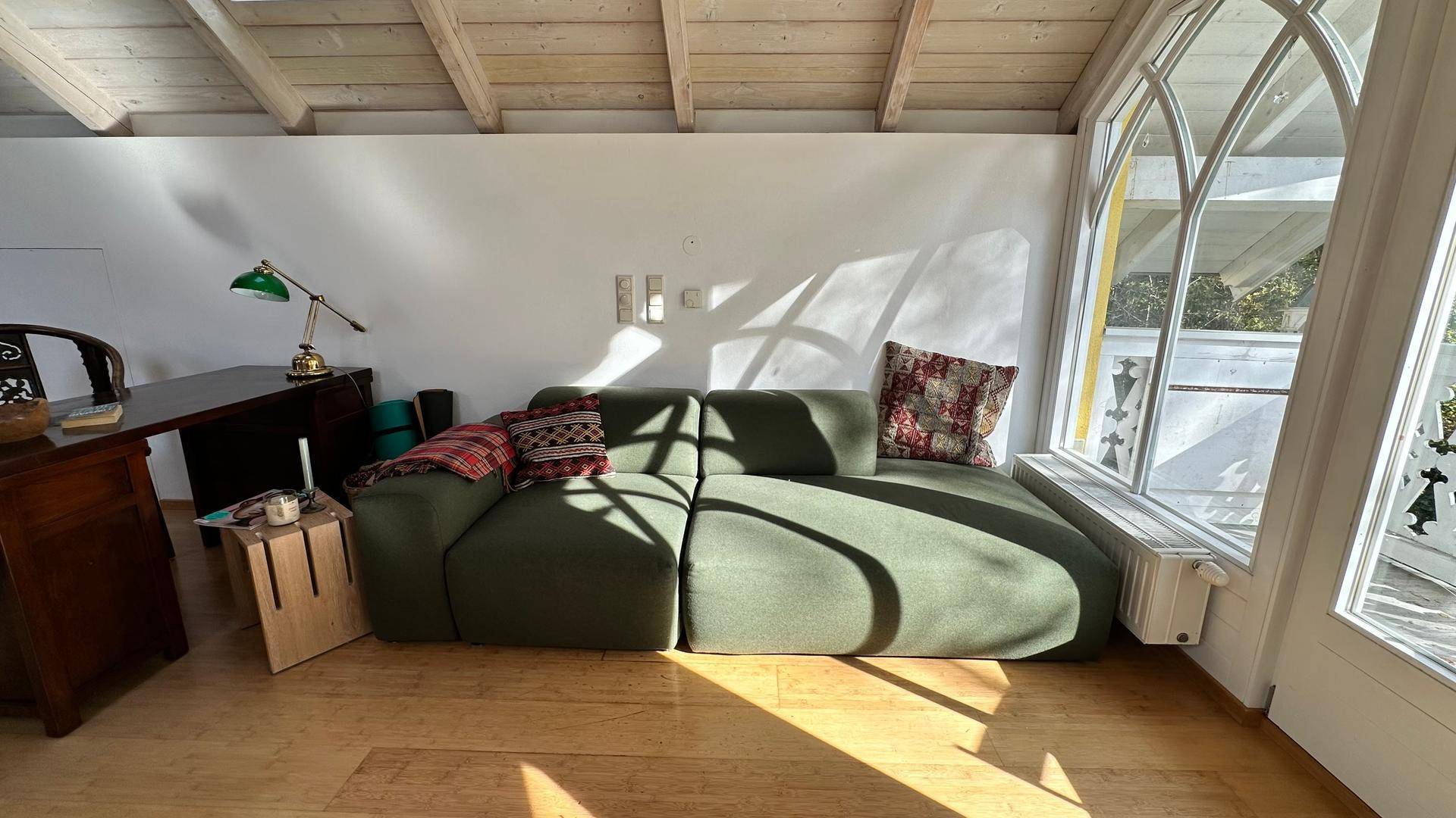 Heller Wohnbereich mit einem großen grünen Sofa, Holzbalkendecke und großen Bogenfenstern mit Blick ins Grüne.