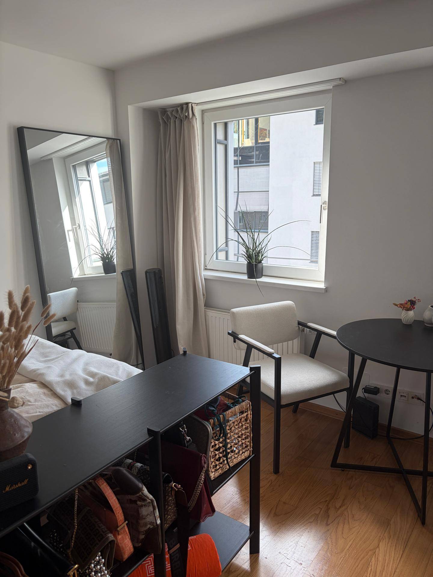 Helles Schlafzimmer mit großem Fenster, einem bequemen Stuhl, kleinem Tisch und einem hohen Spiegel.
