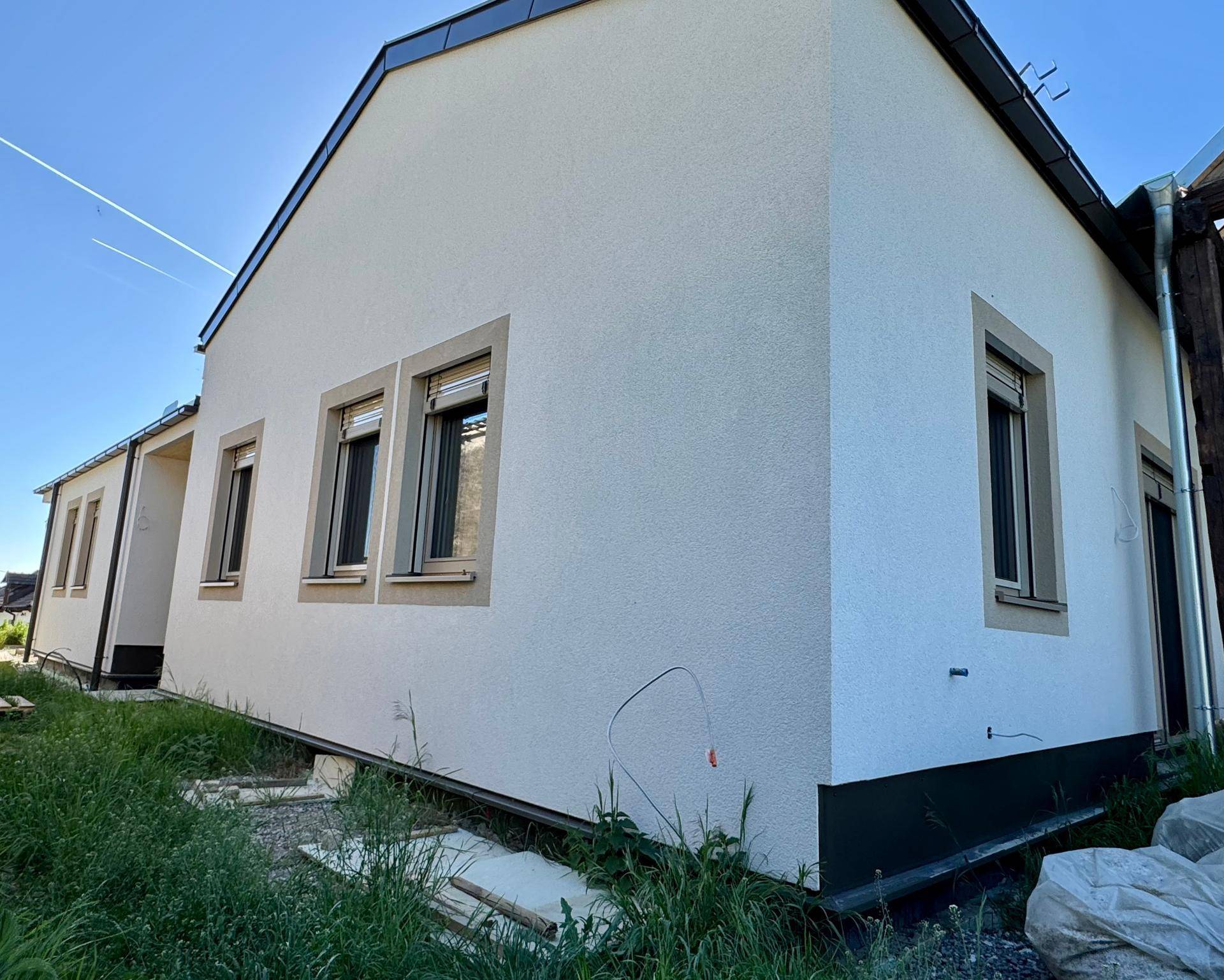 Seitenansicht eines Neubaus mit weißer Putzfassade, braunen Fensterrahmen und Rollläden im Bauzustand.