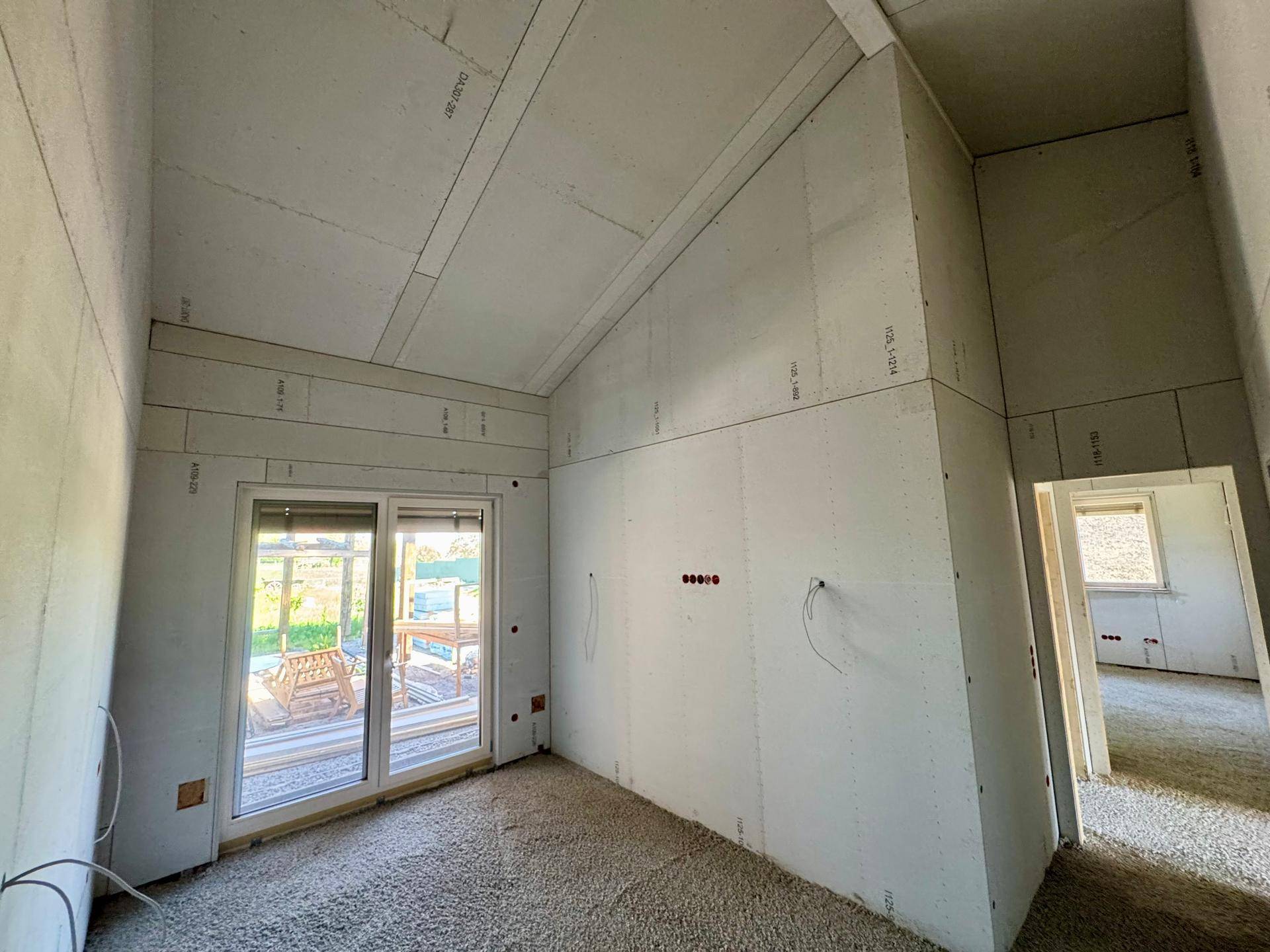 Zimmer im Rohbau mit Dachschräge, Schiebetür zum Außenbereich und Blick ins Grüne.