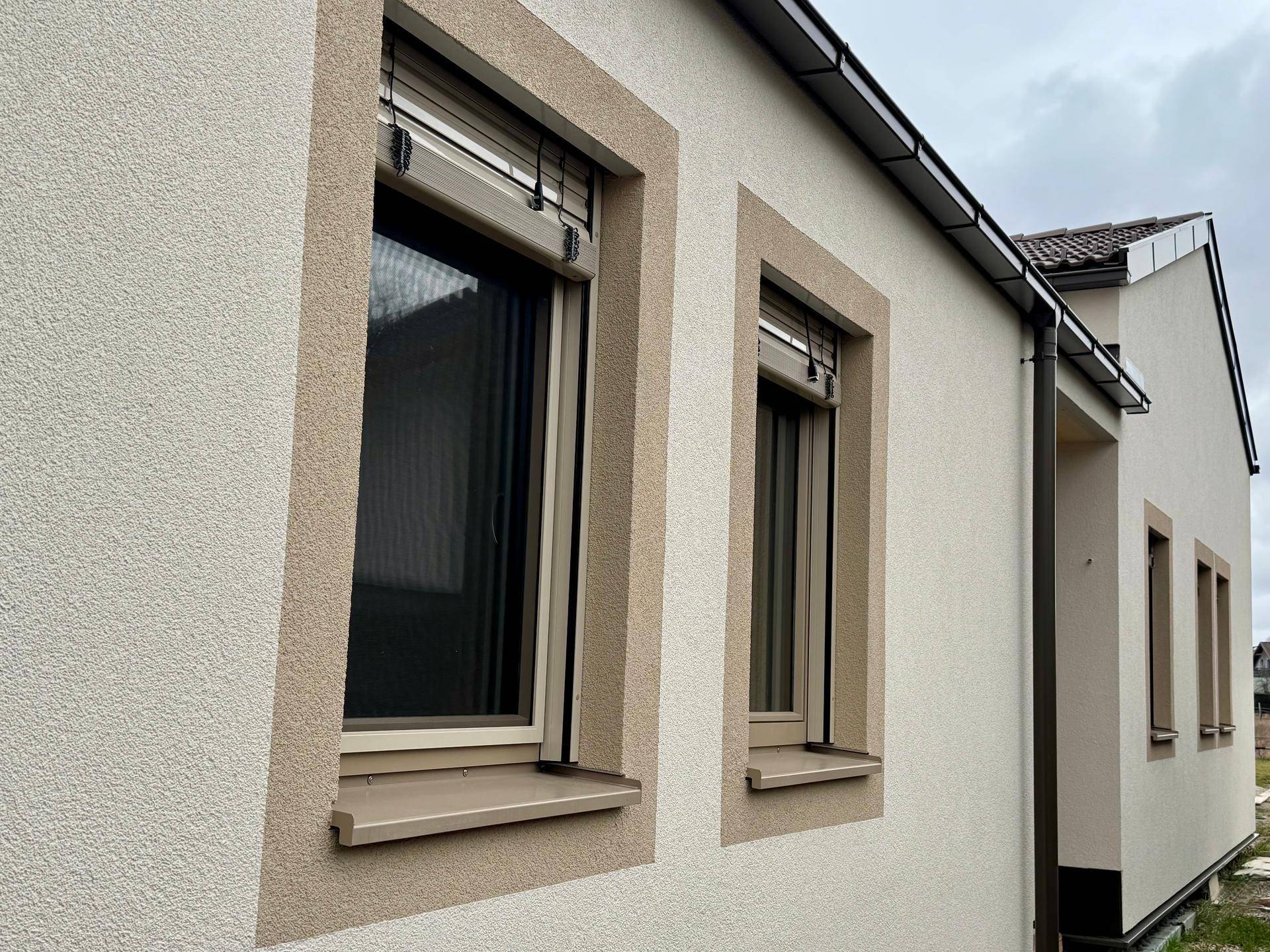 Fassade eines Neubaus mit beigen Fensterrahmen und Rollläden unter einem bewölkten Himmel.