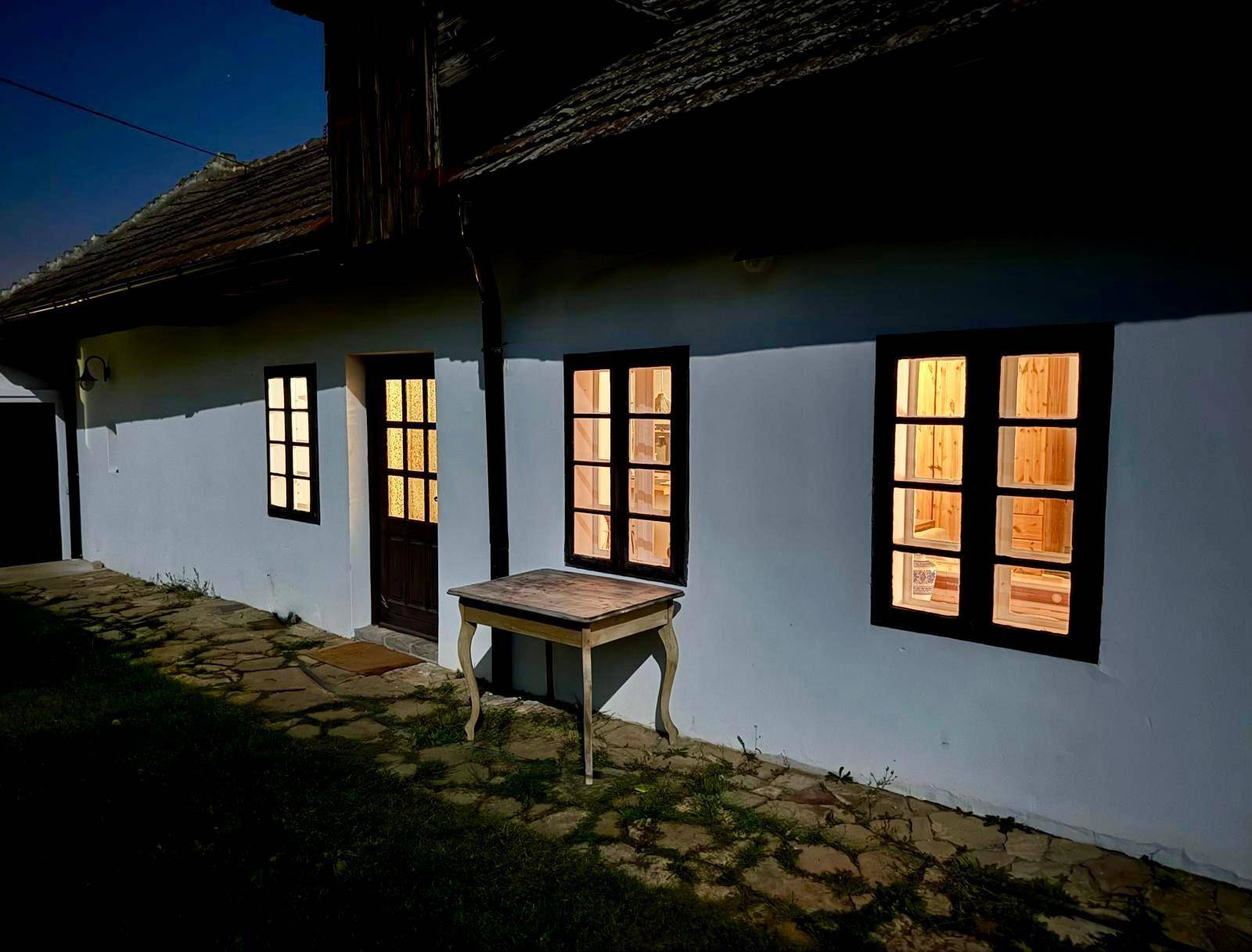 Altes Bauernhaus bei Nacht mit beleuchteten Fenstern und rustikalem Holztisch vor der Fassade.