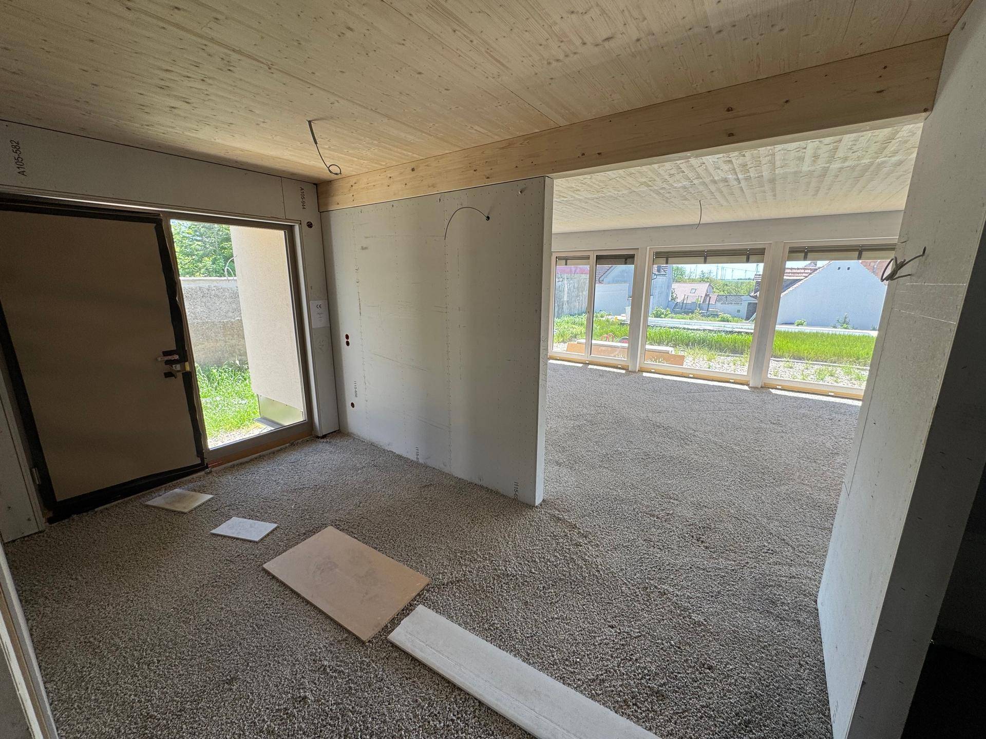 Eingangsbereich im Rohbau mit Holzbalkendecke und Blick durch große Fenster ins Grüne.