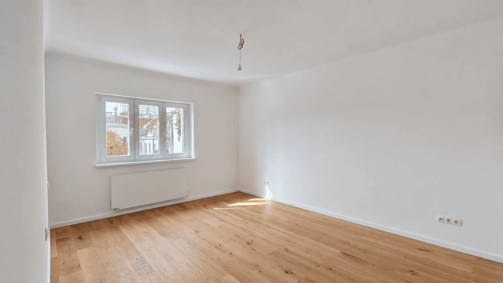 Heller, leerer Raum mit Holzboden und einem Fenster, das den Blick nach draußen freigibt.