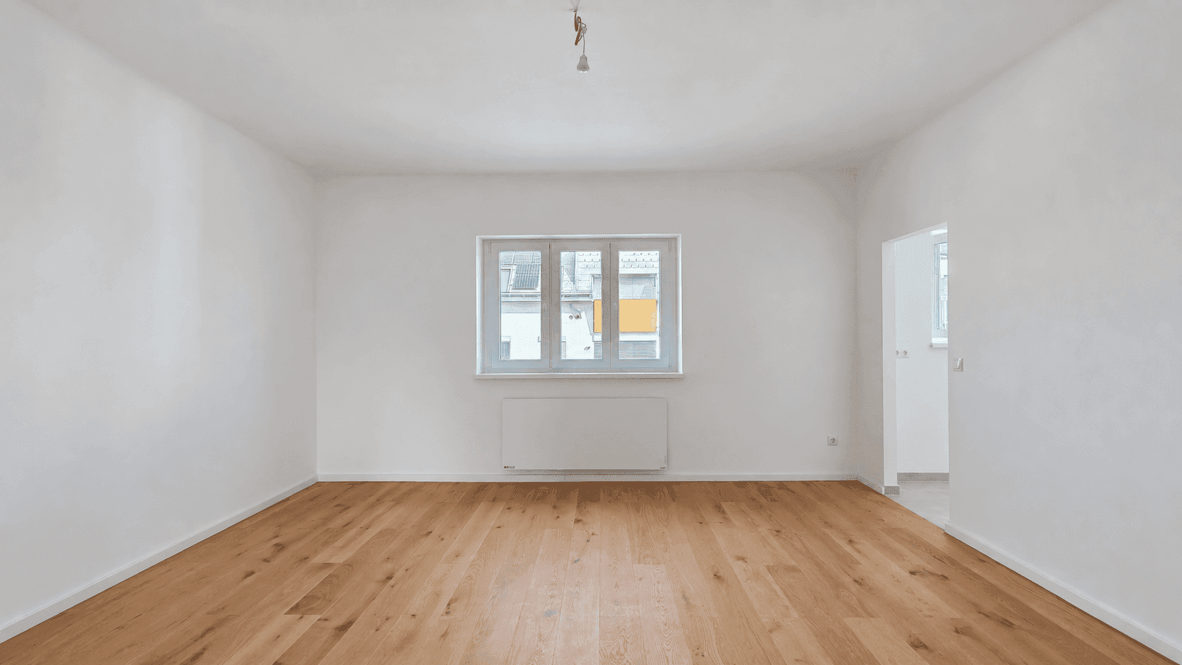 Leerer Raum mit hellem Holzboden, weißen Wänden und einem Fenster mit Heizkörper darunter.