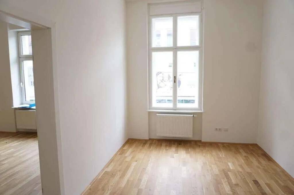 Heller Raum mit Holzboden und großem Fenster, mit Blick in einen angrenzenden Bereich.