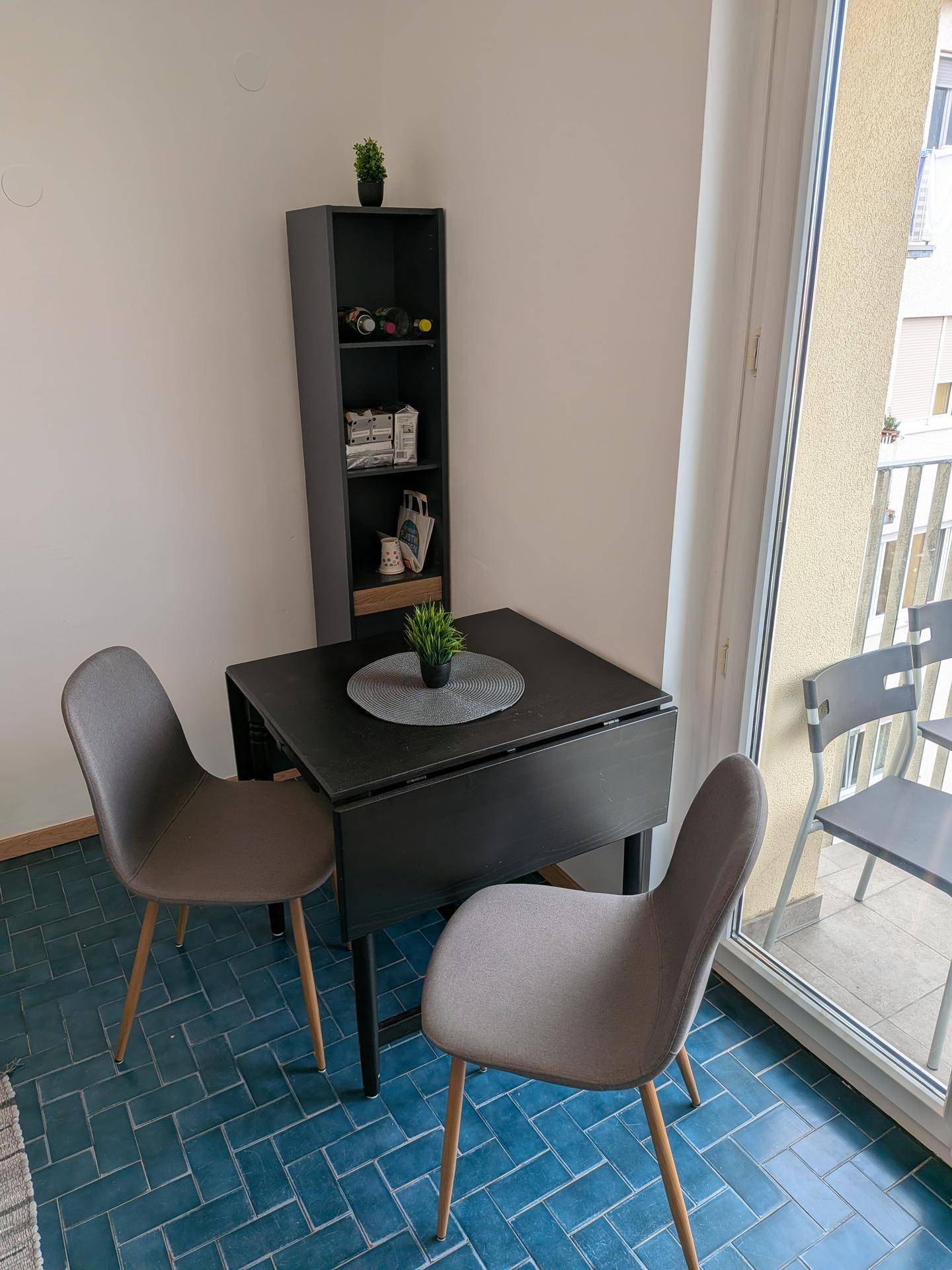 Essbereich in der Küche mit klappbarem Tisch, Stühlen und Regal, auf blauen Fliesen.