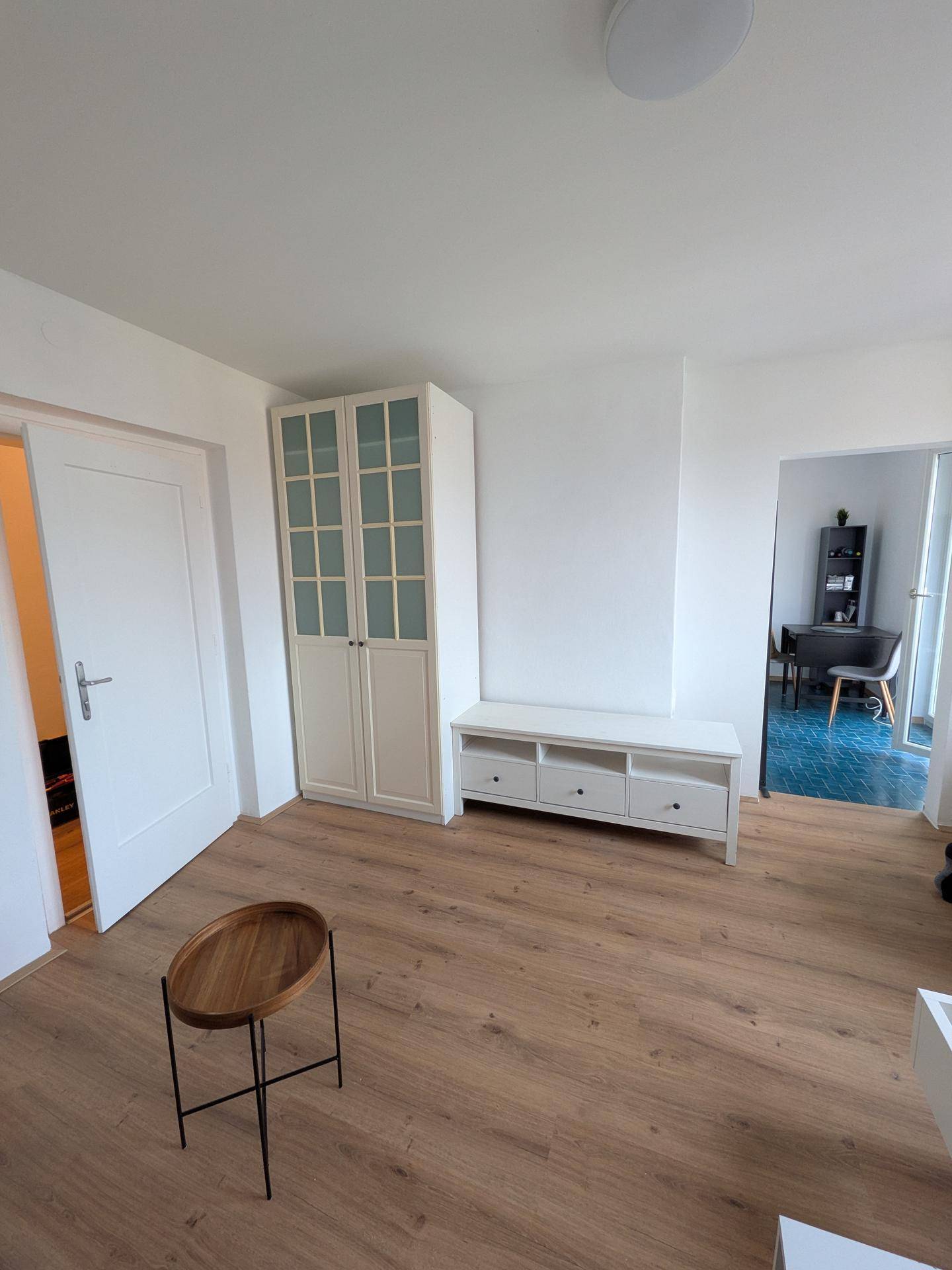 Wohnbereich mit hellem Laminatboden, weißem Schrank und offenem Übergang zur Küche.