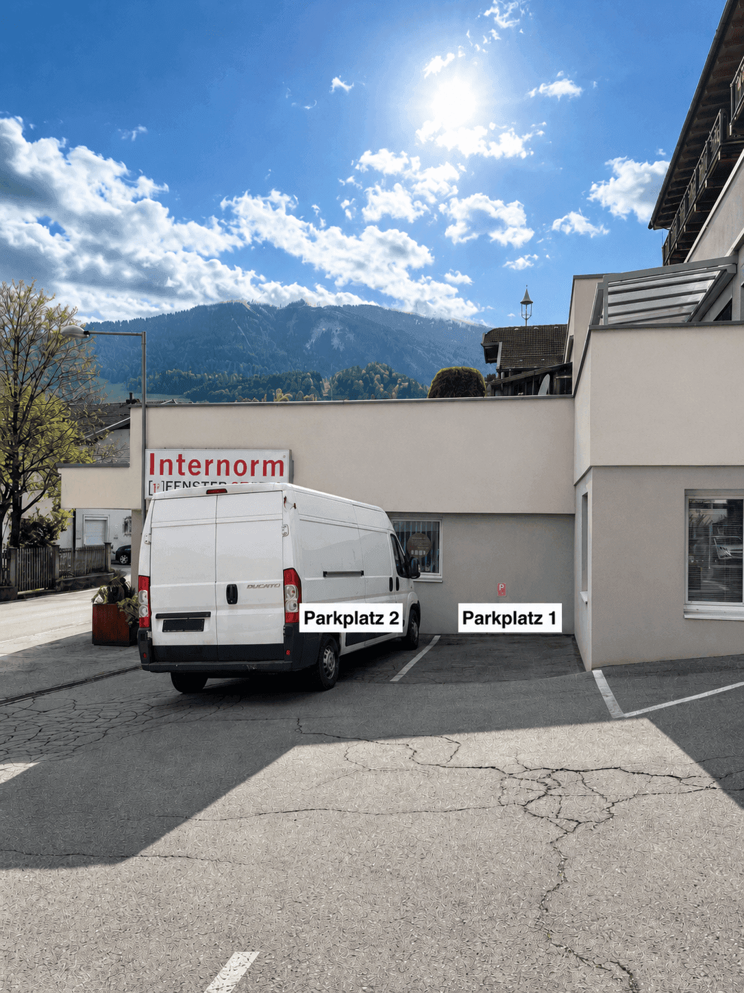 Außenansicht des Gebäudes mit Parkplätzen und einem Lieferwagen unter strahlendem Sonnenschein und Bergen im Hintergrund.