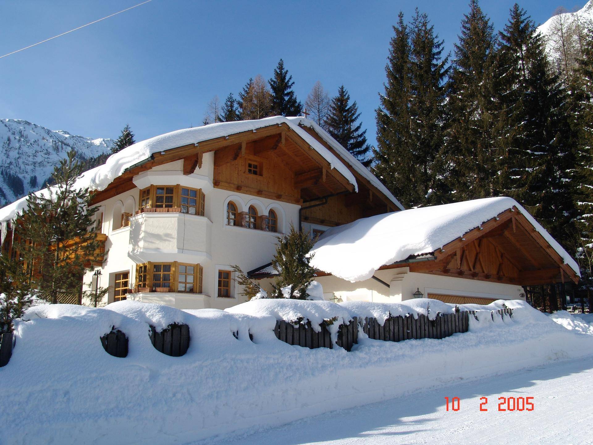 Traditionelles Haus im Winter, bedeckt mit Schnee, umgeben von Nadelbäumen und Bergen.