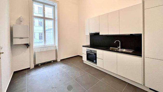 Moderne Küche mit weißen Schränken, schwarzer Arbeitsplatte und integriertem Backofen, hell durch ein Fenster.