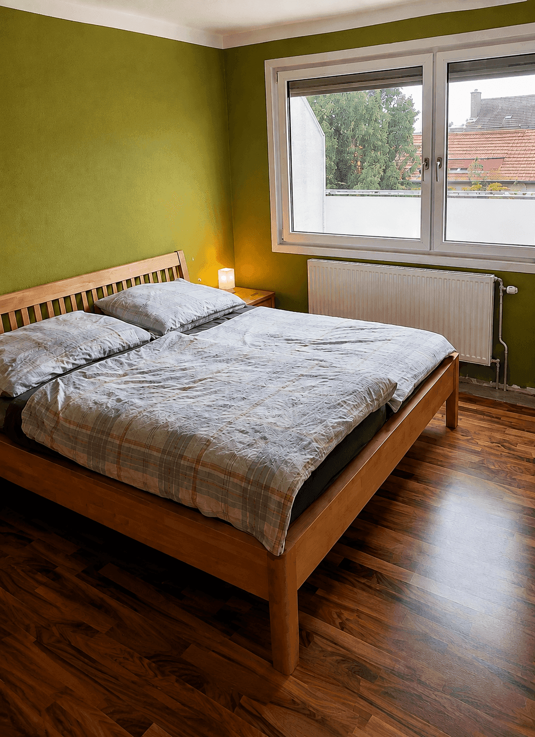 Schlafzimmer mit Holzbett, grünen Wänden und Fenster, das viel Tageslicht hereinlässt.