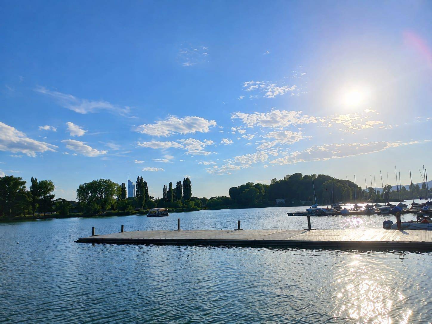 Idyllischer Seeblick mit Bootssteg, Yachthafen und Stadtkulisse am Horizont bei Sonnenuntergang.