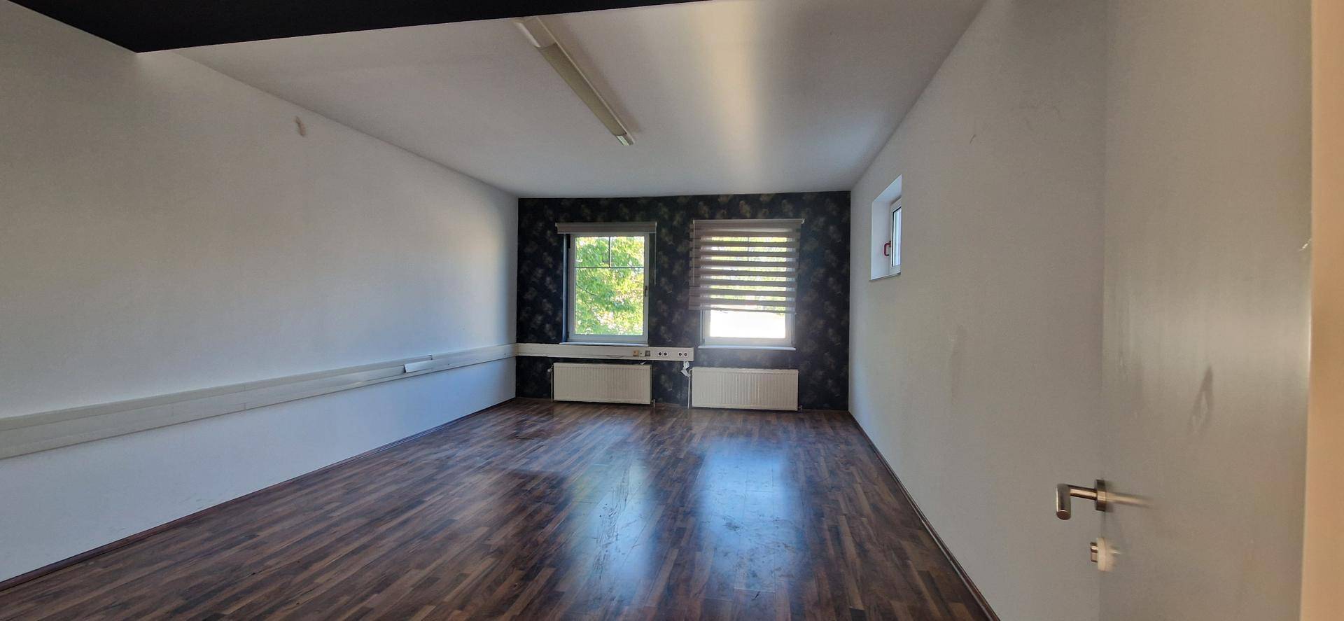 Leerer Büroraum mit dunklem Laminatboden, weißen Wänden und zwei Fenstern mit Jalousien, die für Privatsphäre sorgen.