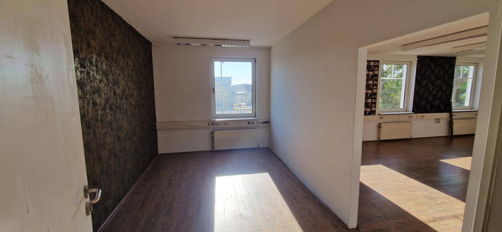 Helles Büro mit dunkler Mustertapete an einer Wand, Laminatboden und einem Fenster, das Tageslicht spendet.