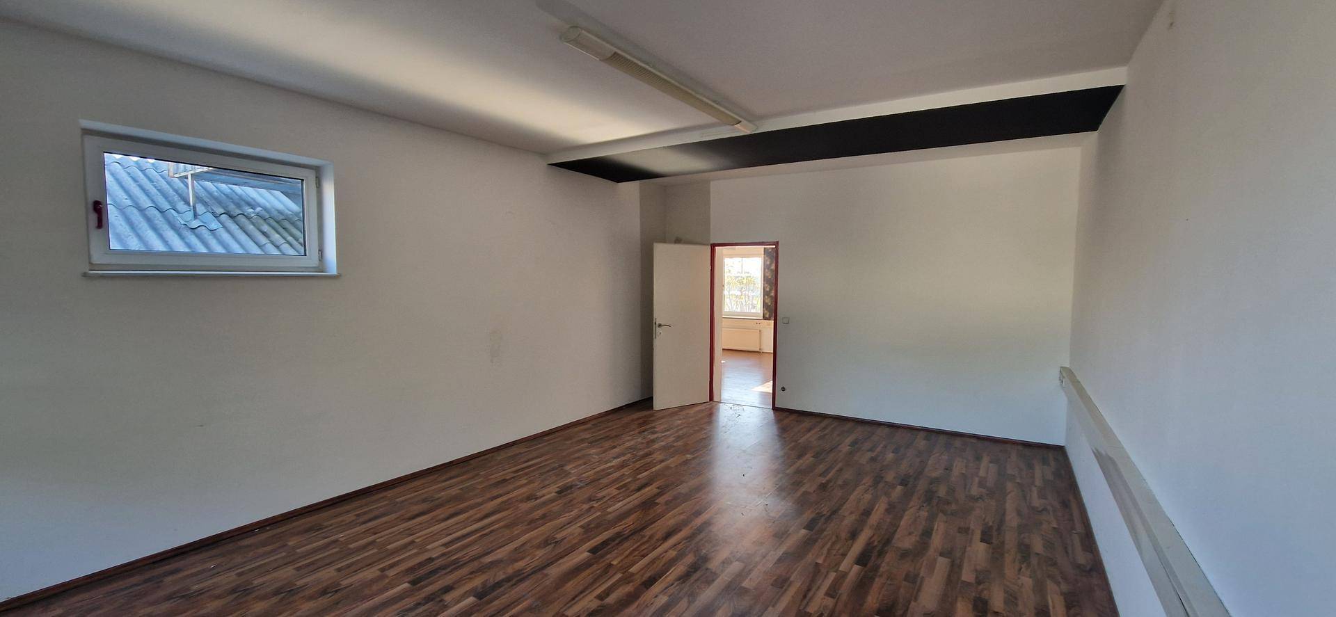 Helles Büro mit dunklem Laminatboden, weißen Wänden und einem Fenster mit Blick auf ein Dach, das Tageslicht hereinlässt.