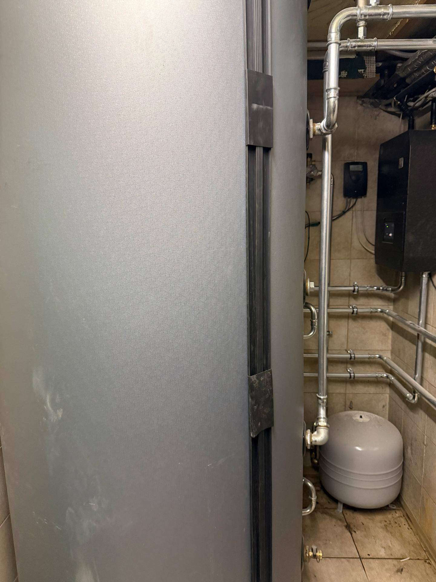 Kellerraum mit großem Warmwasserspeicher, sichtbaren Rohren und gefliestem Boden.