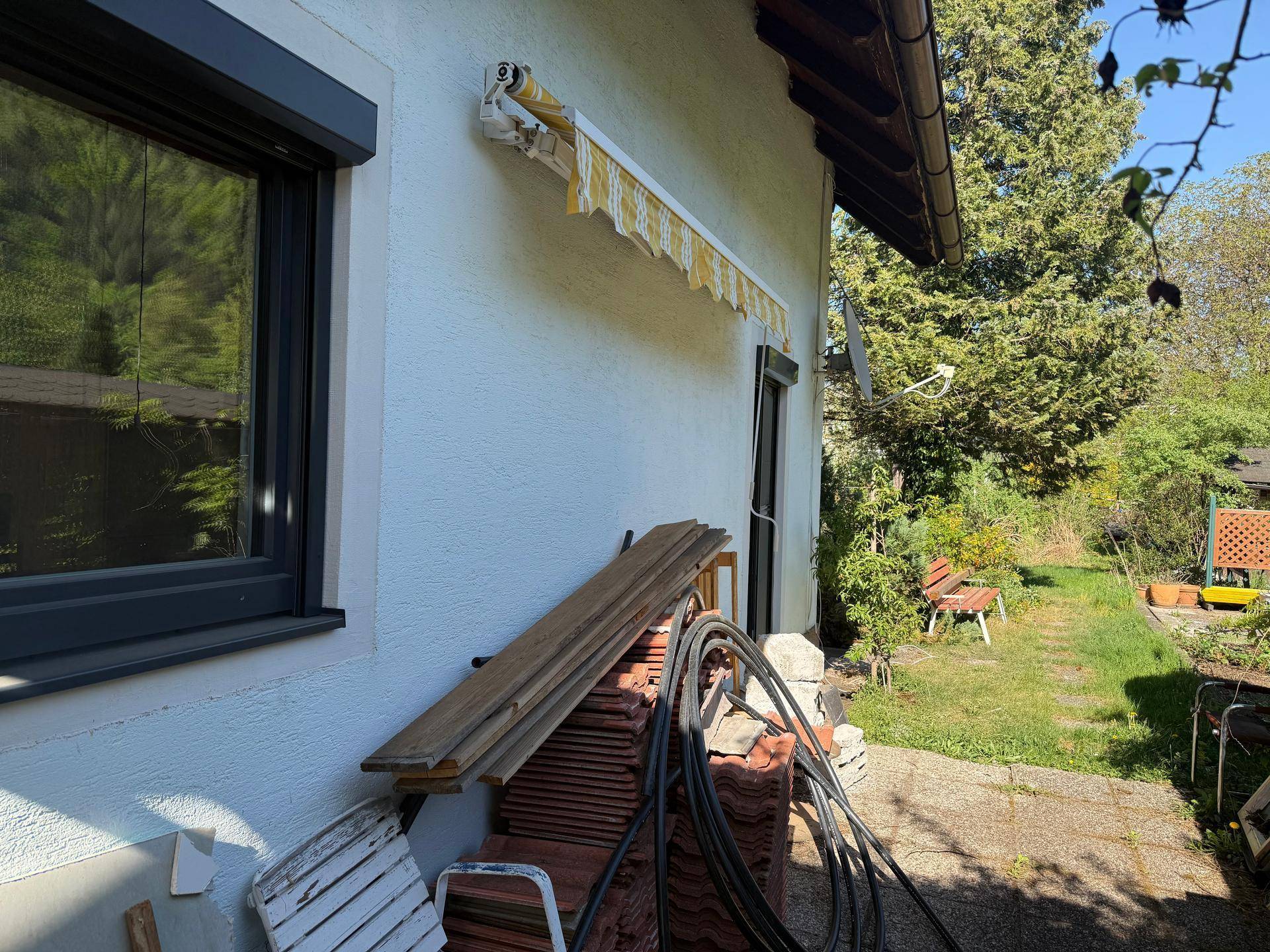 Seitenansicht des Hauses mit kleiner gepflasterter Fläche, Markise und Gartenbereich.