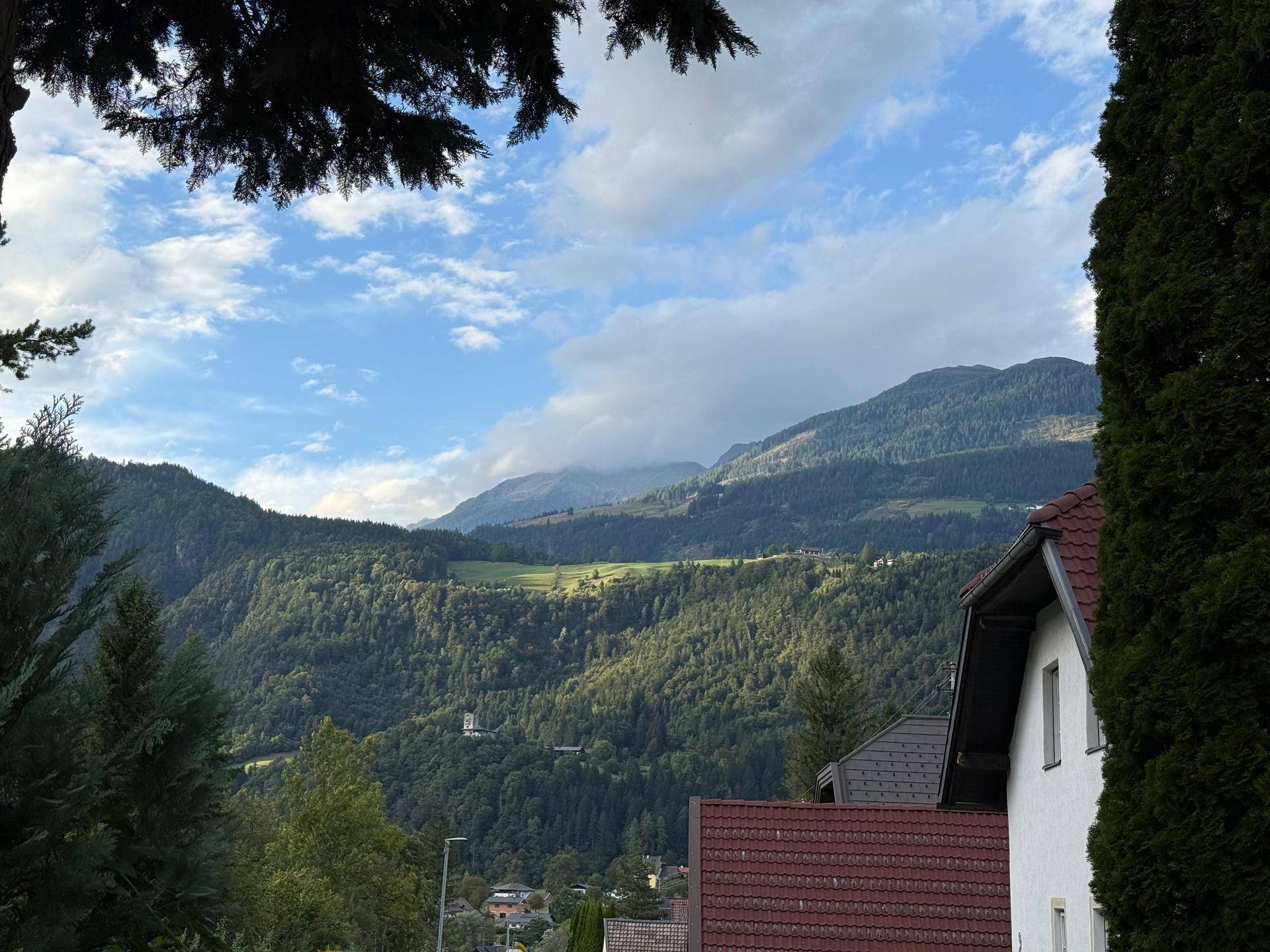 Malerischer Blick auf bewaldete Berge und grüne Hügel mit einem Hausdach im Vordergrund.