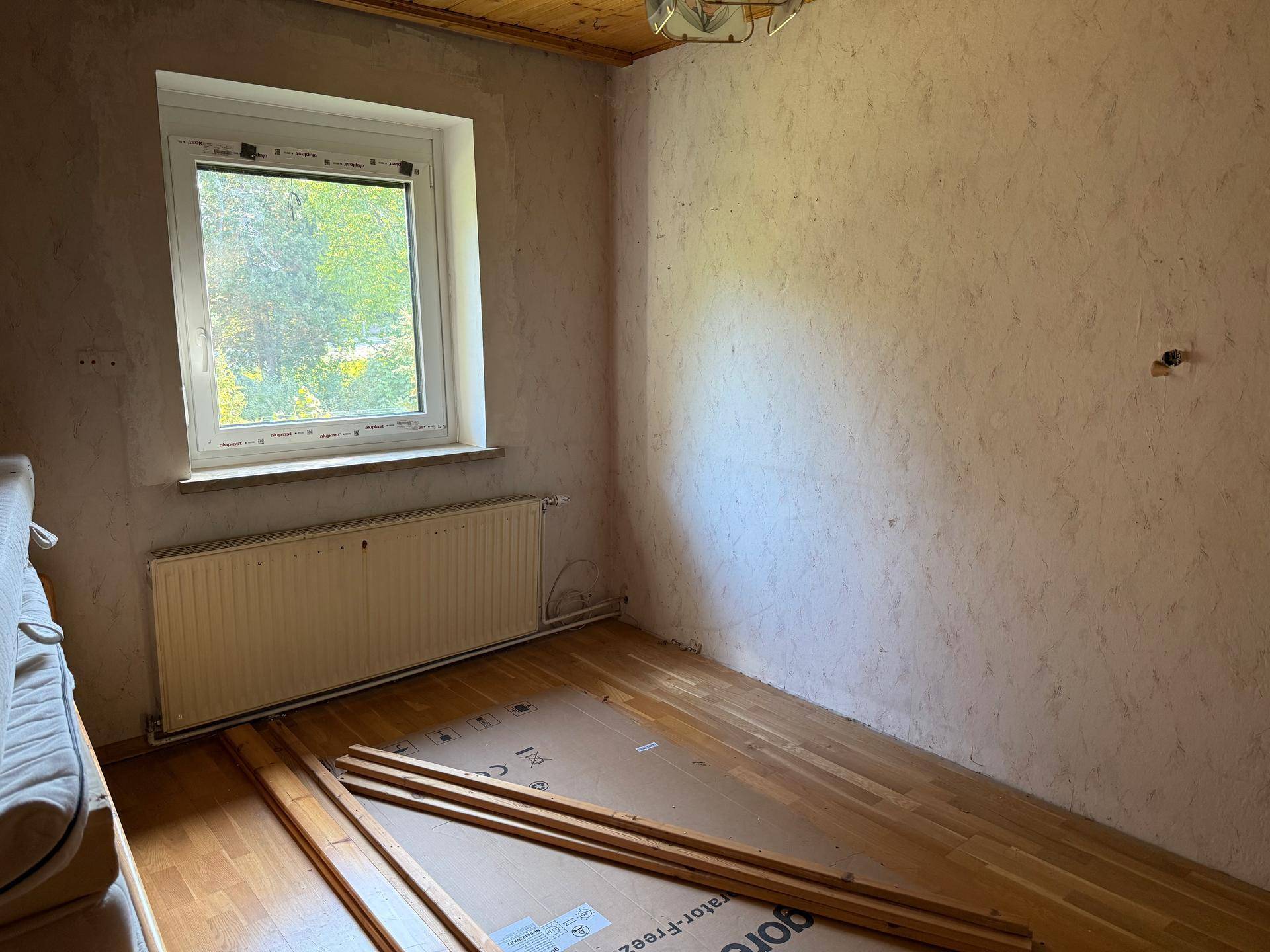 Renovierungsbedürftiges Zimmer mit unfertigen Wänden, Holzboden und einem Fenster mit Blick ins Grüne.